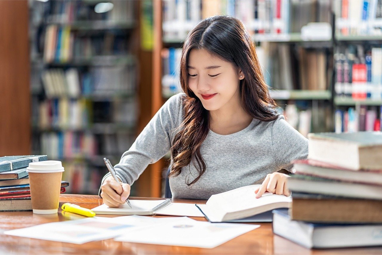 A woman is sitting at a library, taking notes from a book.